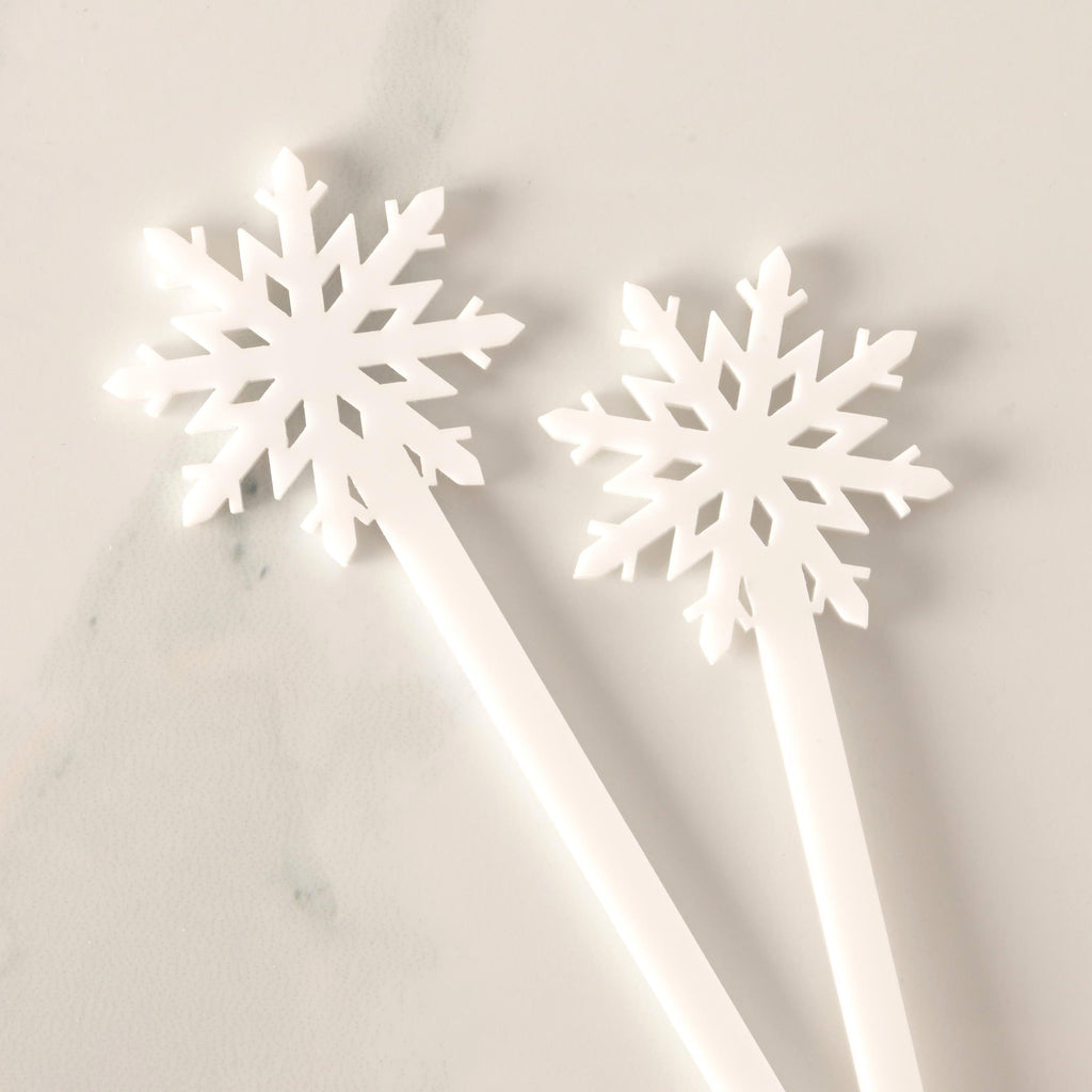 A close-up of two white snowflake-topped cocktail stirrers, overlapping slightly. The stirrers have a clean, solid white appearance and are displayed on a white marble surface.