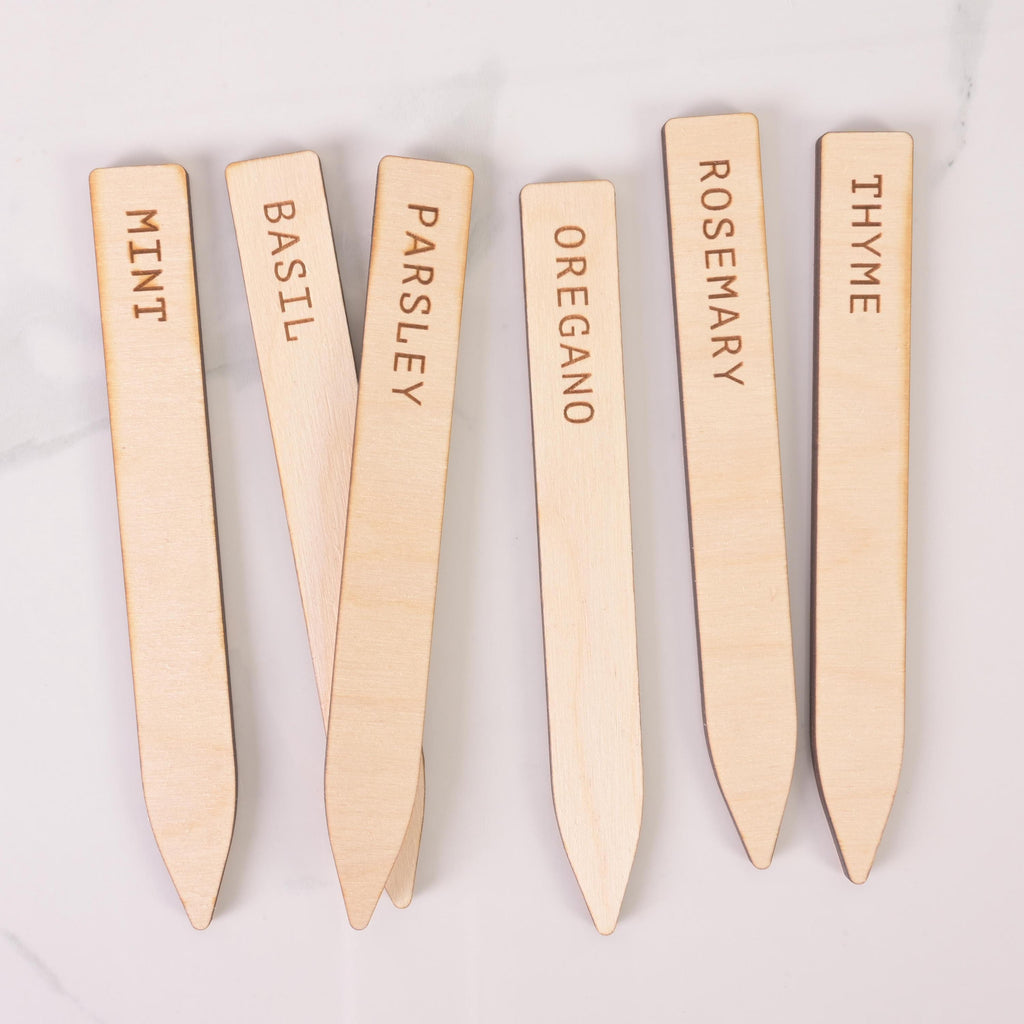 Six engraved wooden herb markers labeled Mint, Basil, Parsley, Oregano, Rosemary, and Thyme, arranged on a flat surface for gardening use.
