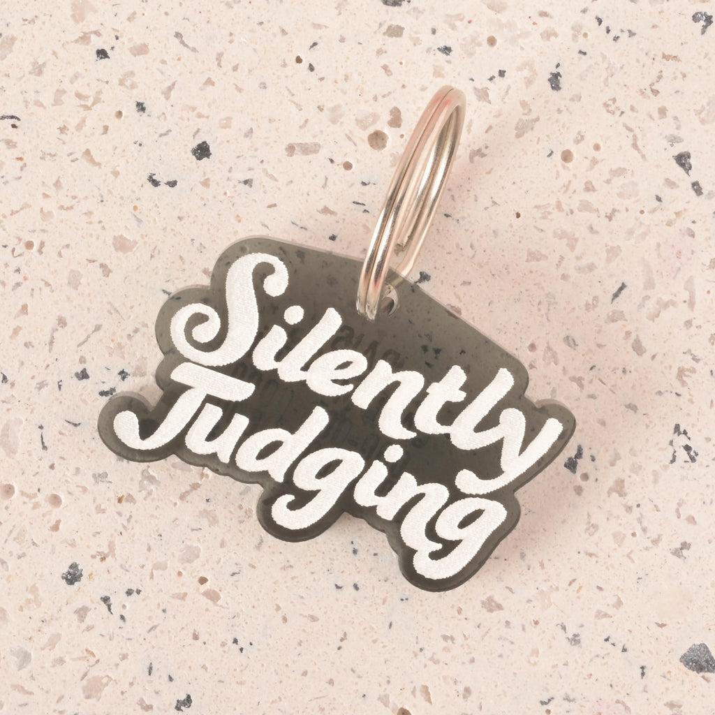 A close-up shot of a black pet tag in the shape of a speech bubble that says "Silently Judging" in a white groovy font. The tag is attached to a silver ring and is resting on a light-colored, speckled surface.