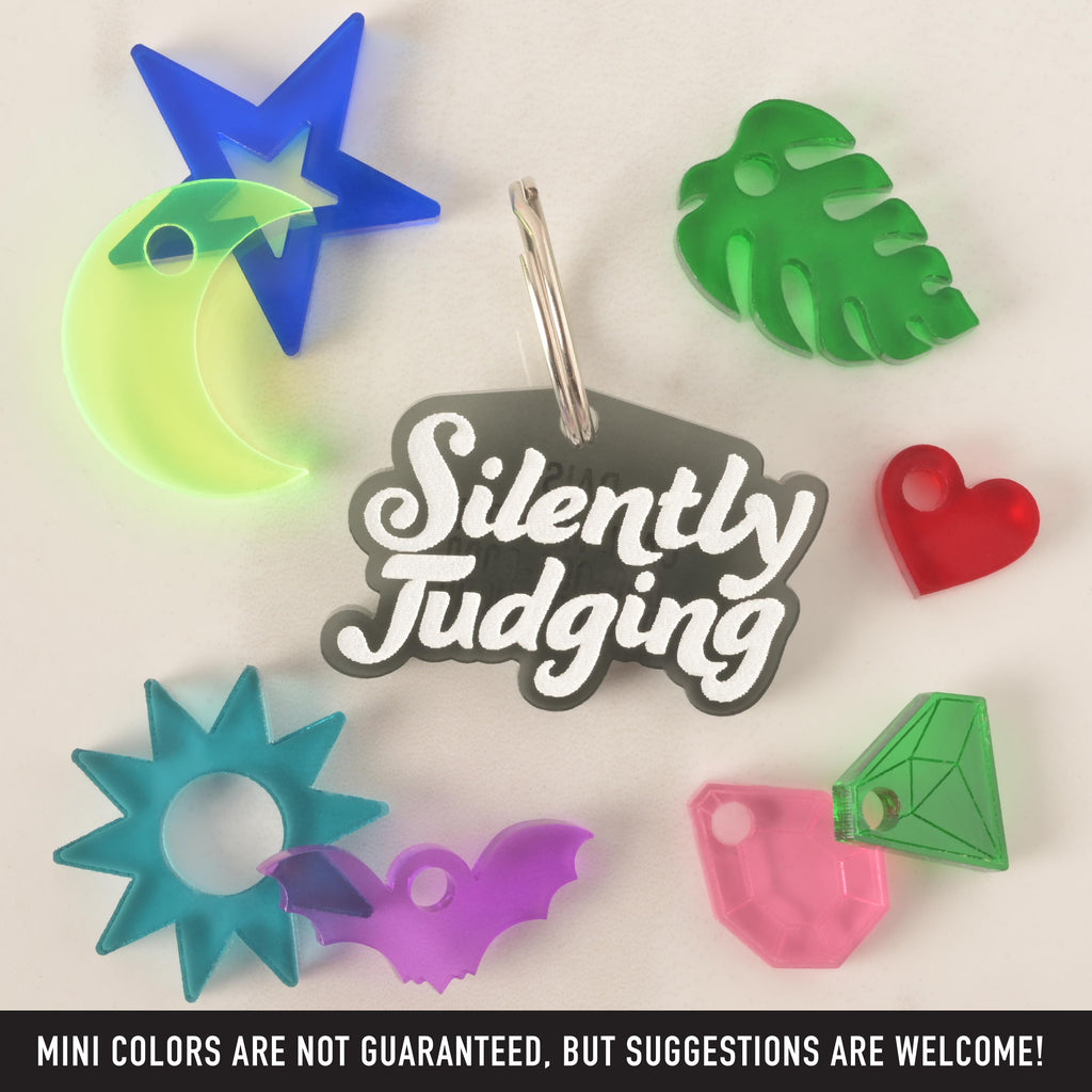 A collection of pet tags and charms scattered on a white surface. The central piece is a black "Silently Judging" tag in a white groovy font. Surrounding it are various charms, including a neon yellow moon with a blue star, a green monstera leaf, a red heart, a blue sun, a purple bat, and two different colored gemstones. The image also contains the text "MINI COLORS ARE NOT GUARANTEED, BUT SUGGESTIONS ARE WELCOME!"