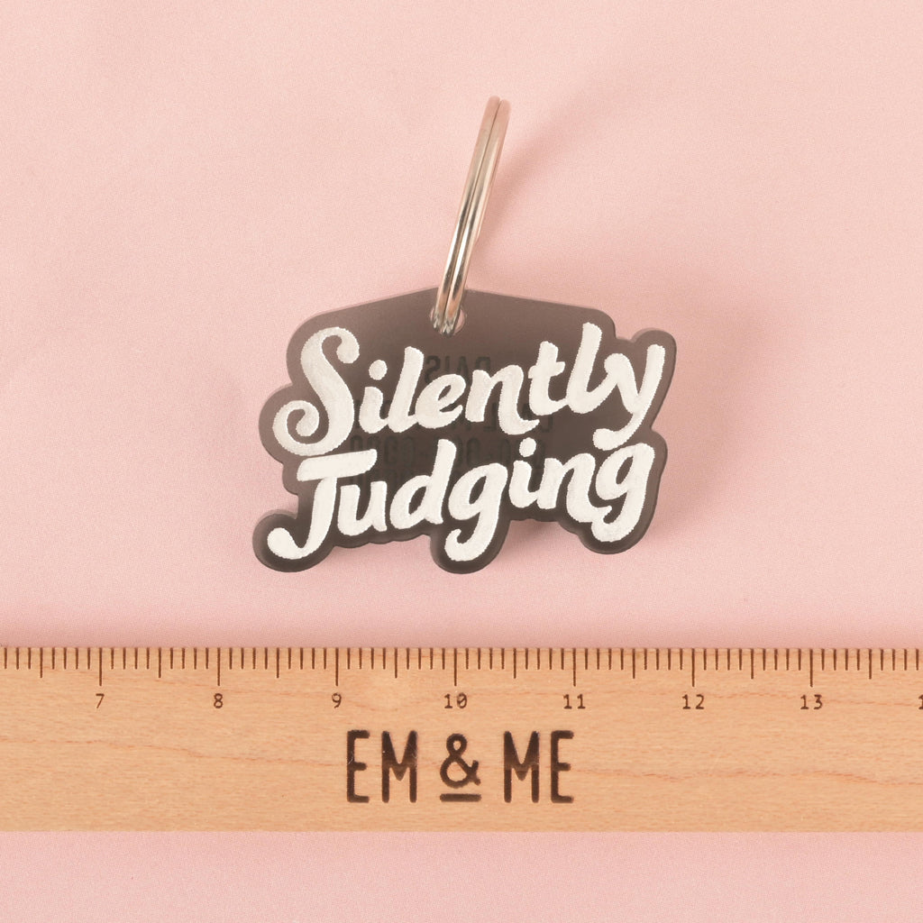 A transparent gray pet tag in the shape of a speech bubble that says "Silently Judging" in a white groovy font. A ruler with the "EM & ME" logo is placed below the tag to show its size. The tag is shown on a solid pink background.