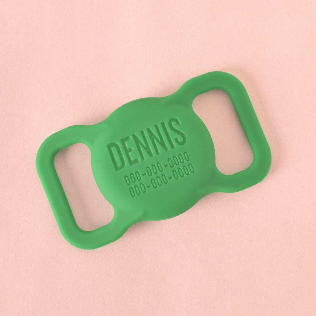 A close-up shot of a custom, silent pet ID tag designed to slide onto a collar. The tag is a bright green and is engraved with the name "DENNIS" and a phone number. It is displayed on a solid pink background.