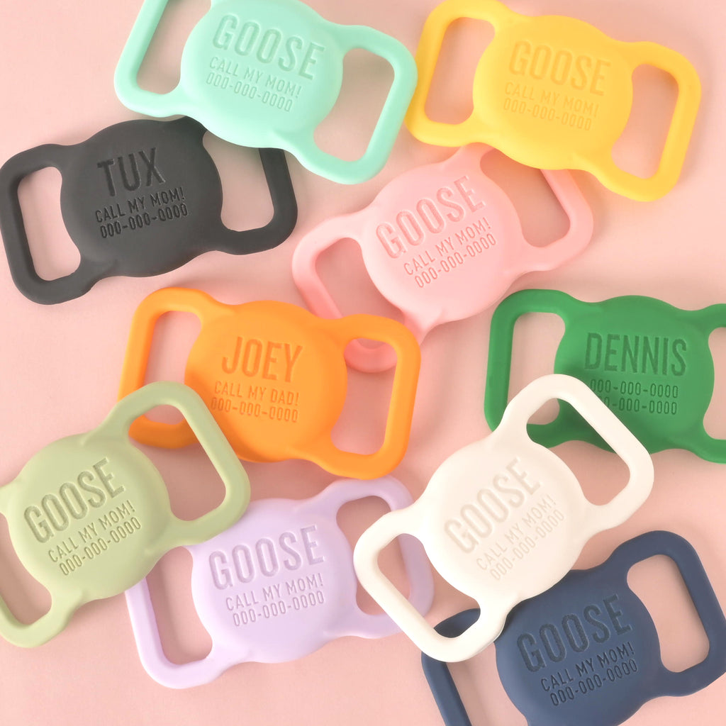 A scattered group of custom, silent, slide-on pet ID tags in various colors, including mint green, yellow, pink, sage green, light purple, charcoal gray, white, dark blue, and orange. The tags are engraved with different names like "GOOSE," "TUX," "JOEY," and "DENNIS," along with funny phrases like "CALL MY MOM!" or "CALL MY DAD!", and a phone number. The tags are displayed on a solid pink background.