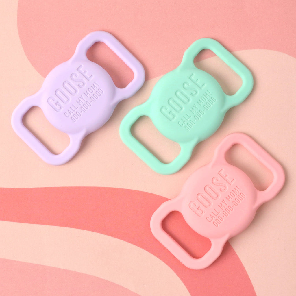 A group of three custom, silent pet ID tags, all engraved with "GOOSE," "CALL MY MOM!", and a phone number. The tags are in pastel colors: light lavender, mint green, and light pink. They are designed to slide onto a collar and are displayed on a light pink and reddish-brown wavy-patterned background.