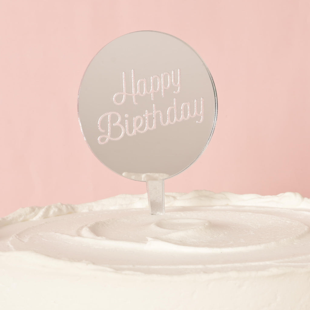 A silver circular mirror cake topper with the words "Happy Birthday" etched into the surface, placed on top of a white frosted cake. The background is a solid pink color.