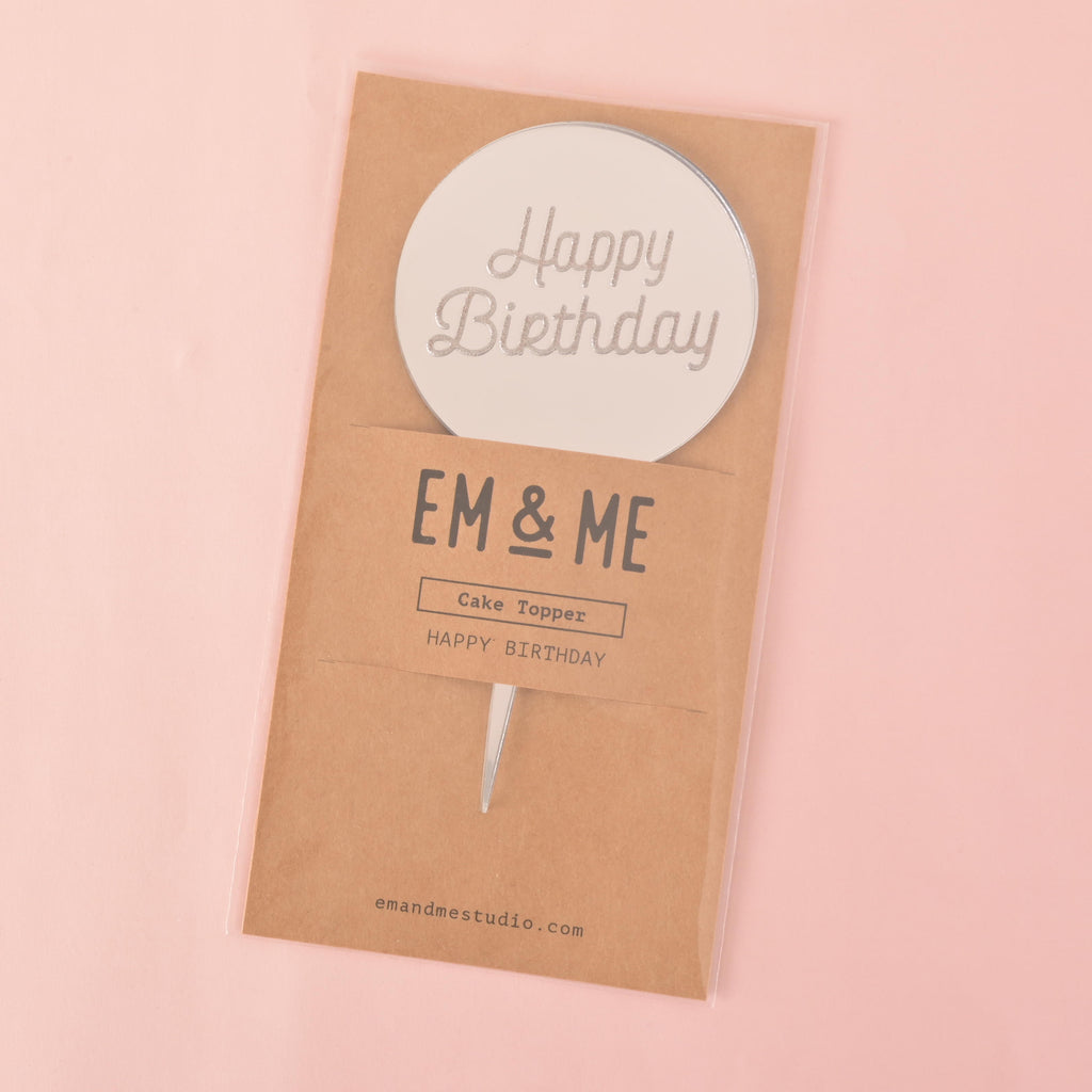 A silver circular mirror cake topper that says "Happy Birthday," sealed in its retail packaging. The brown cardstock packaging has "EM & ME" printed on it. The item is shown on a solid pink background.