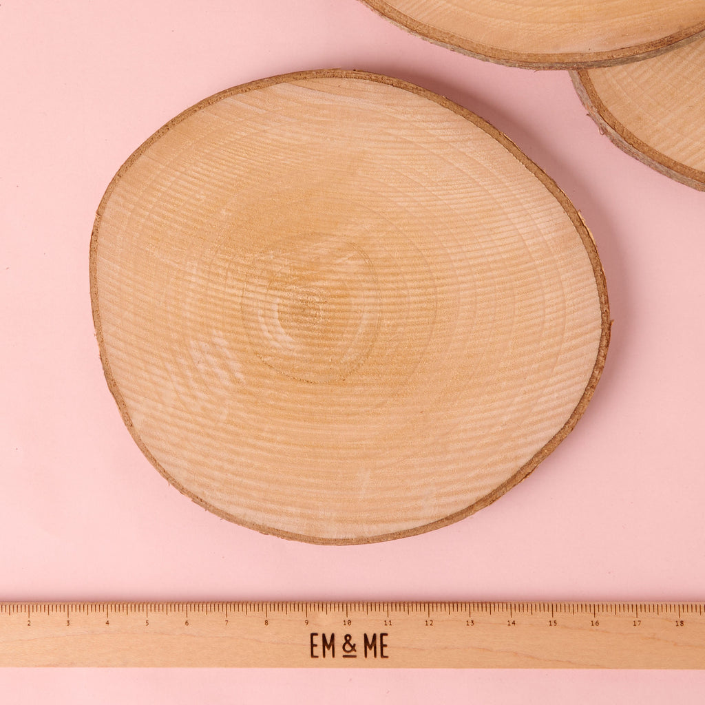 A single circular, natural wood slice with visible growth rings and bark edge, suitable for a coaster or craft project. The disc is displayed above a wooden ruler with the 'EM & ME' logo to show its size.