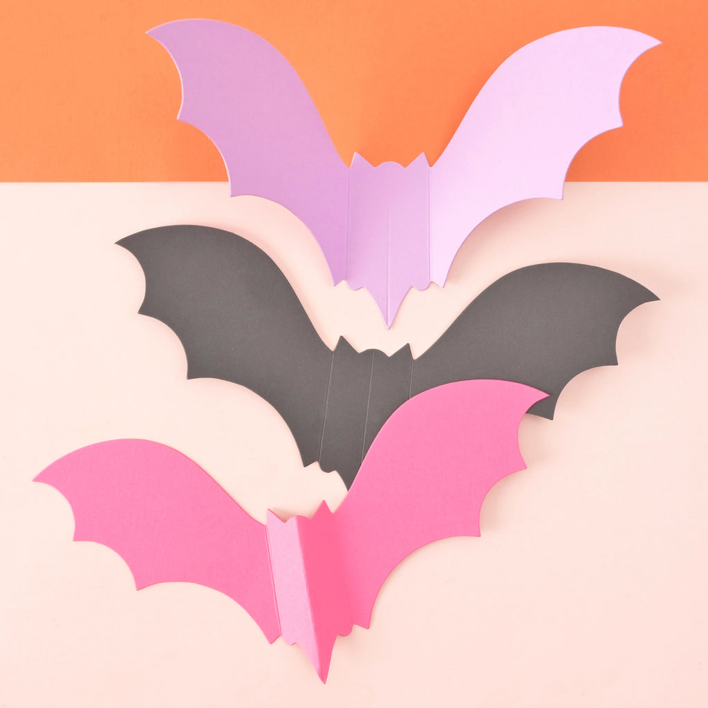 Close up of three single paper bats showing color options of pink, black and lavender on color blocked background