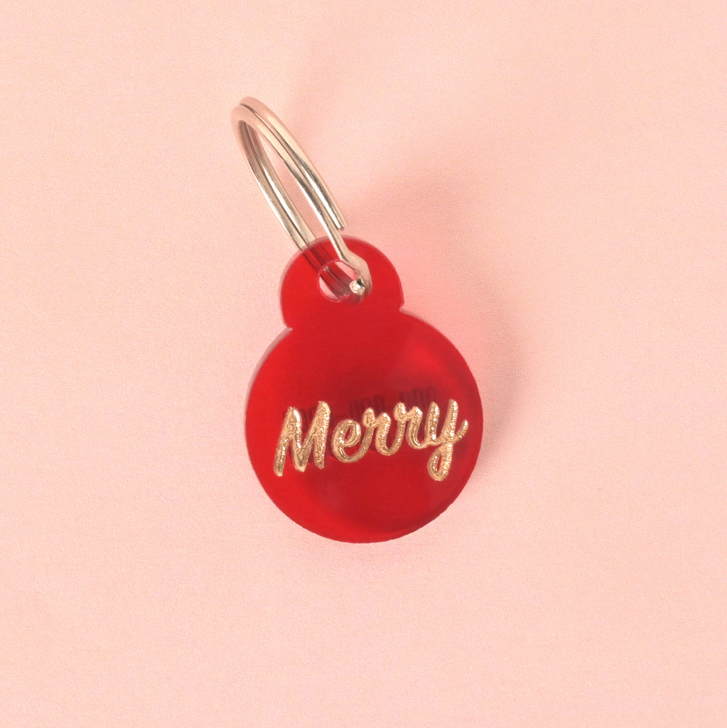 Close-up of a small, red circular pet ID tag shaped like an ornament, featuring the glitter gold slogan 'Merry.' The tag is attached to a silver split ring and displayed on a light pink background.
