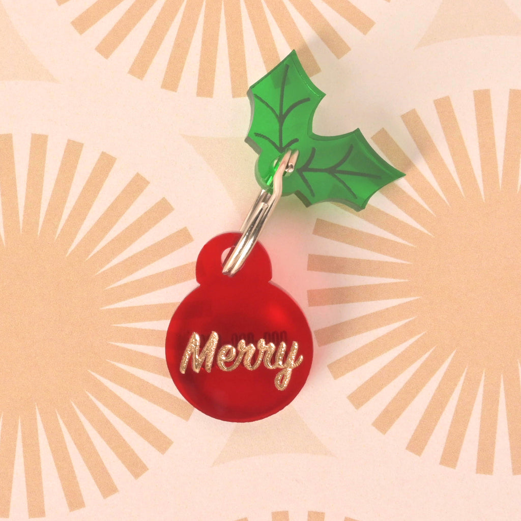 Close-up of a small, red circular pet ID tag with the glitter gold slogan 'Merry.' A green acrylic holly leaf charm is attached above the tag. The tag is displayed on a retro sunburst patterned background.