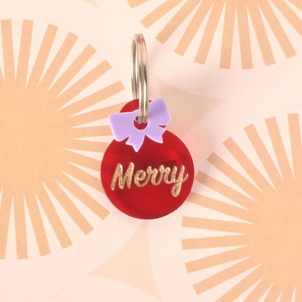 Close-up of a small, red circular pet ID tag with the glitter gold slogan 'Merry.' A light purple (lilac) bow charm is attached above the tag. The tag is displayed on a retro sunburst patterned background.