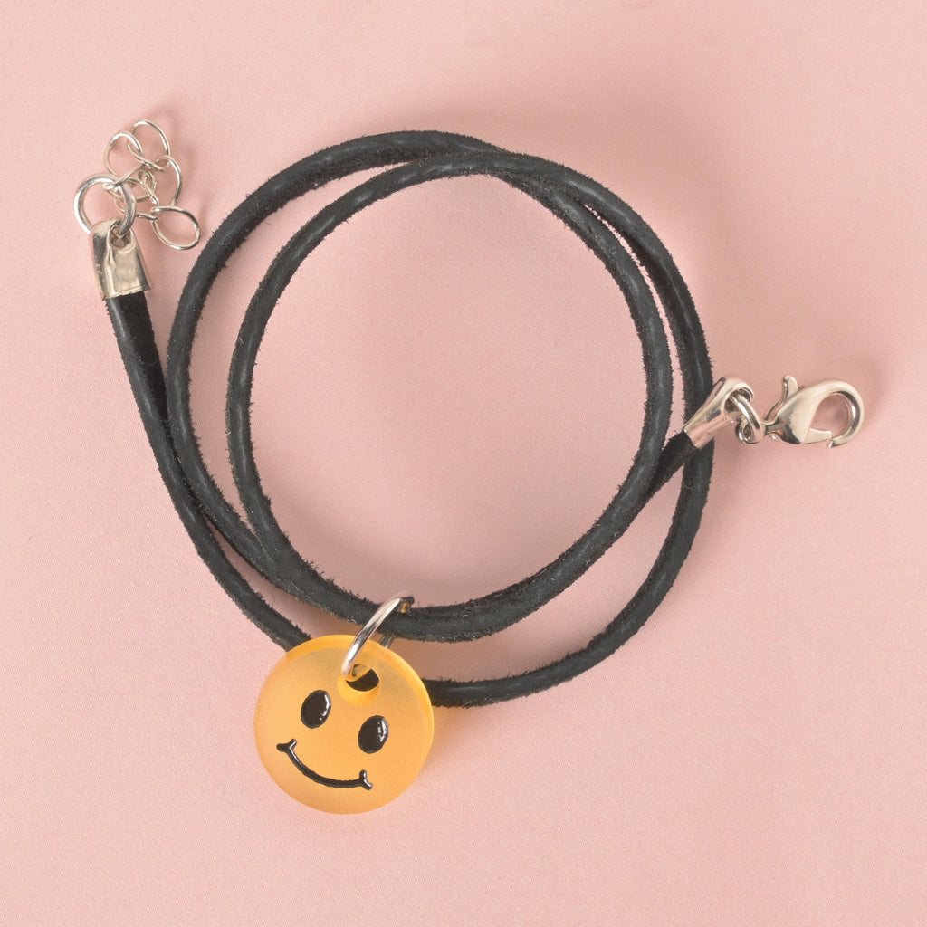 A close-up, top-down view of a translucent orange smiley face charm hanging from a black cord necklace. The necklace has a silver clasp and an extension chain. The charm and necklace are displayed on a solid pink background.