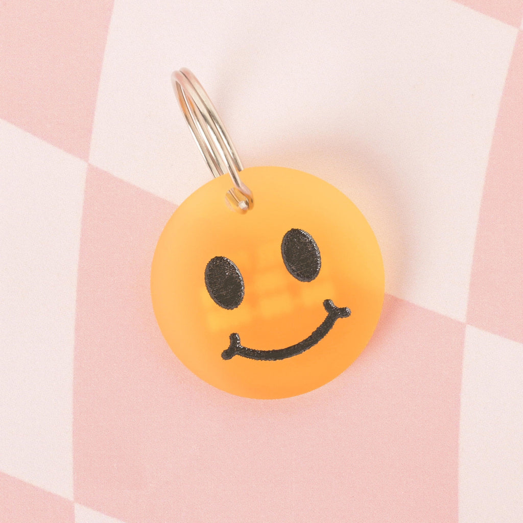 A close-up of a custom, translucent orange, circular pet ID tag with a black smiley face. It is attached to a shiny silver keyring and is displayed on a pink and white checkered background.