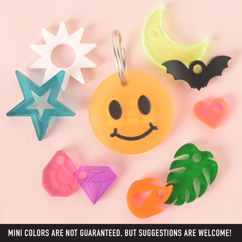 A central, custom yellow smiley face pet tag with a black face and silver keyring. The tag is surrounded by a variety of mini charms, including a turquoise star, a pink gem, a purple gem, an orange flame, a green tropical leaf, a pink heart, a black bat, a glow-in-the-dark crescent moon, and a white sunburst. The items are arranged on a light pink background. A note at the bottom says, "MINI COLORS ARE NOT GUARANTEED, BUT SUGGESTIONS ARE WELCOME!"