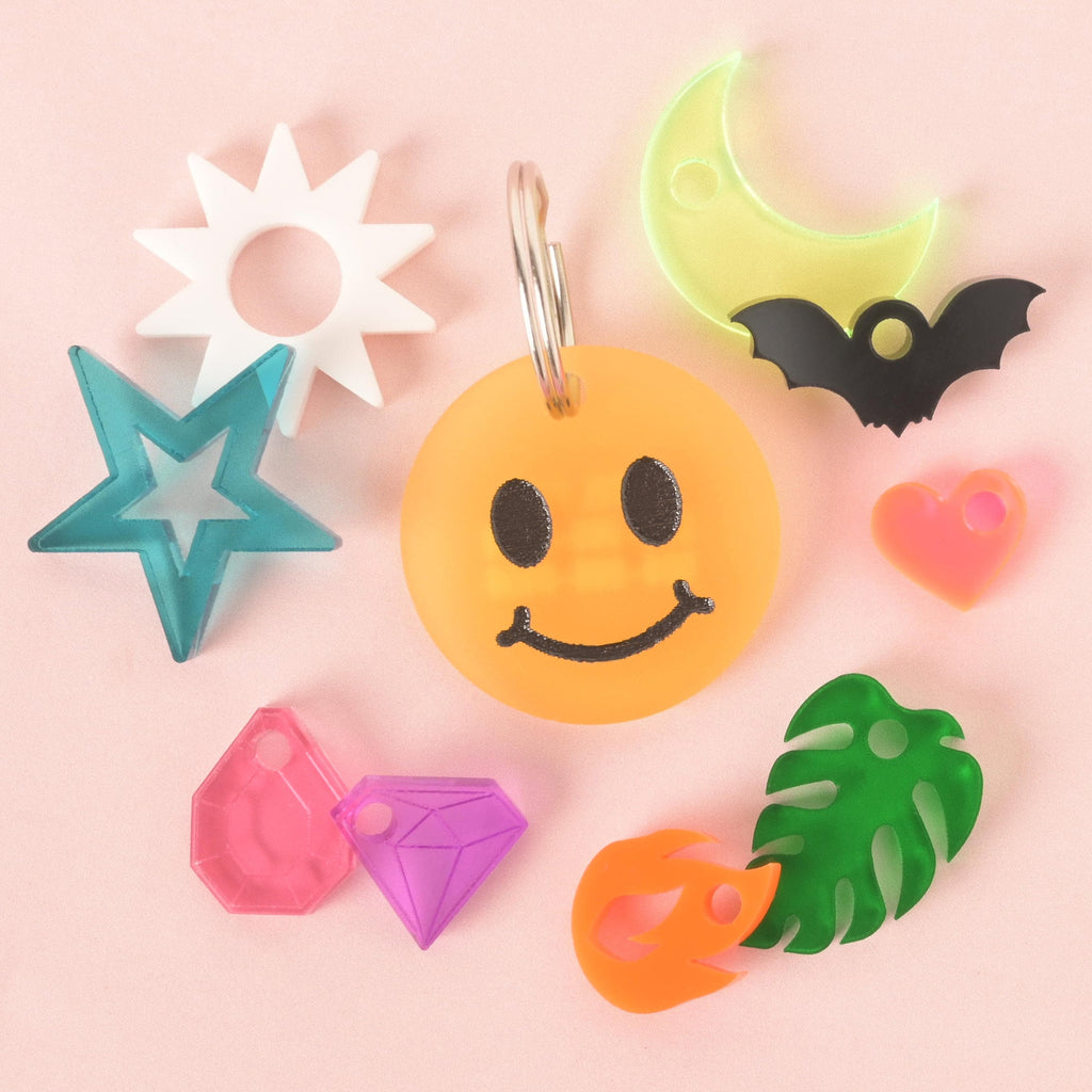 A central, custom orange smiley face pet tag with a black face and silver keyring. The tag is surrounded by a variety of mini charms, including a white sun, a turquoise star, a pink gem, a purple gem, an orange flame, a green tropical leaf, a pink heart, a black bat, and a glow-in-the-dark crescent moon. The items are arranged on a light pink background.