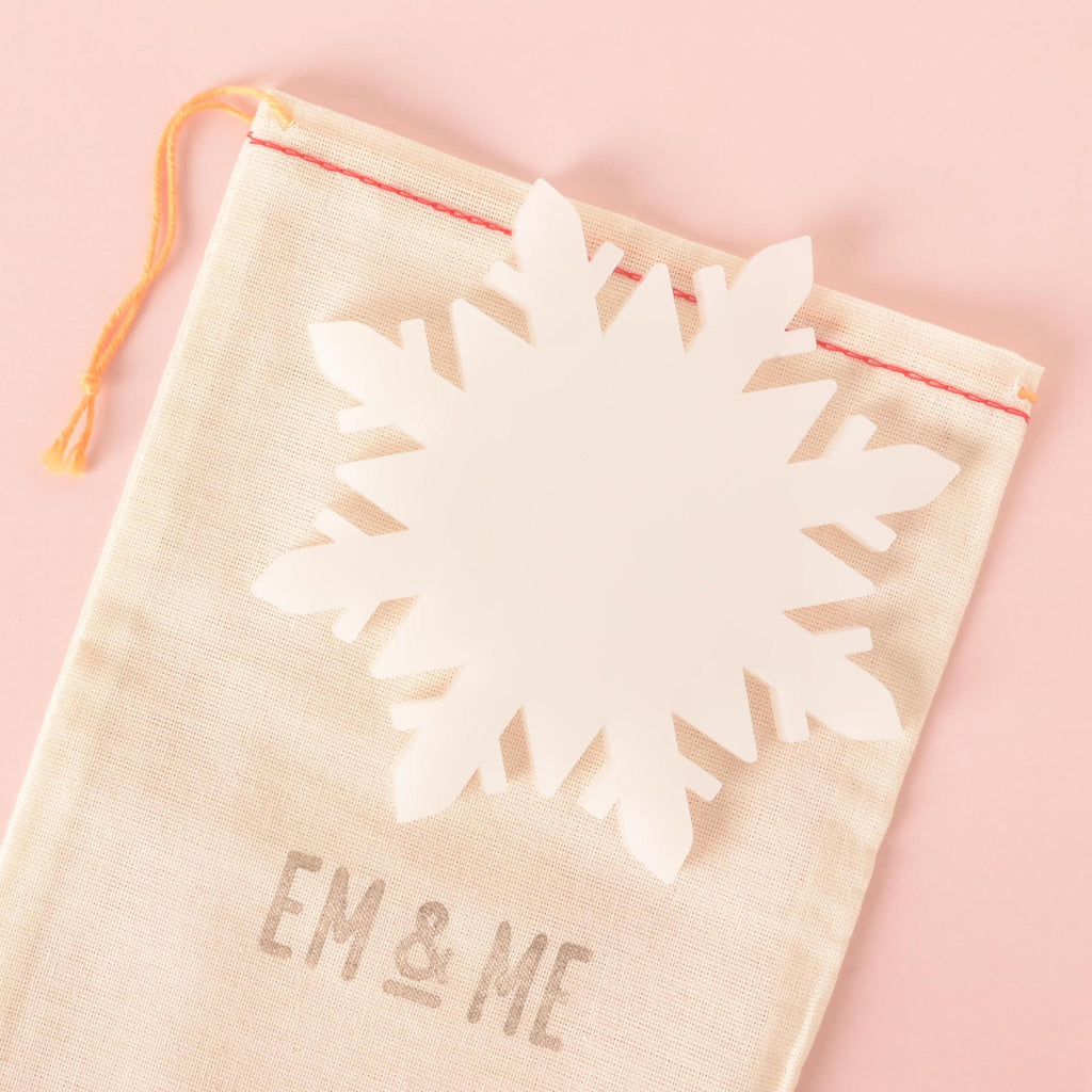 White acrylic snowflake-shaped coaster displayed on a beige drawstring bag with red stitching, perfect for holiday gifting and table protection.