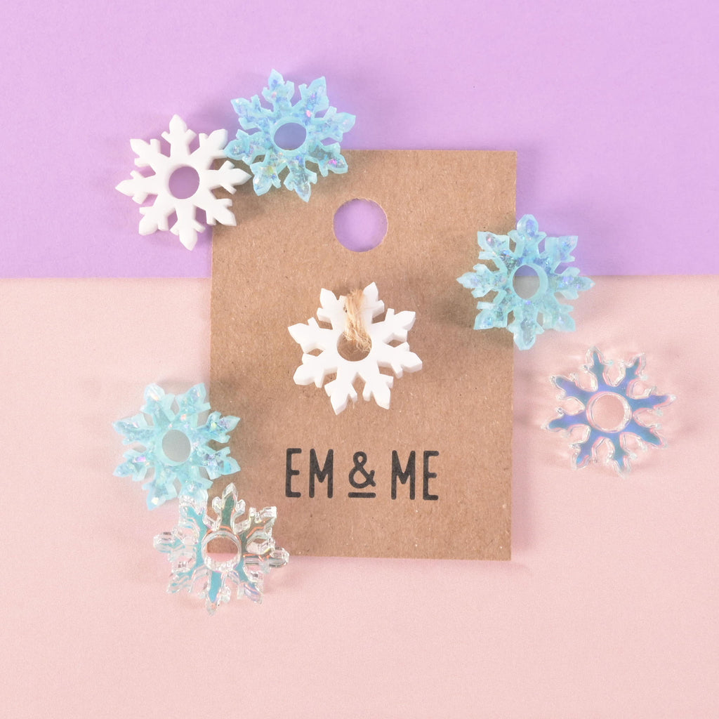 A collection of assorted snowflake-shaped charms and pins. One white snowflake is attached to a brown paper card that reads "EM & ME." Surrounding it are several other snowflakes in various iridescent shades of white and blue. The items are displayed on a background that is half solid purple and half solid pink.