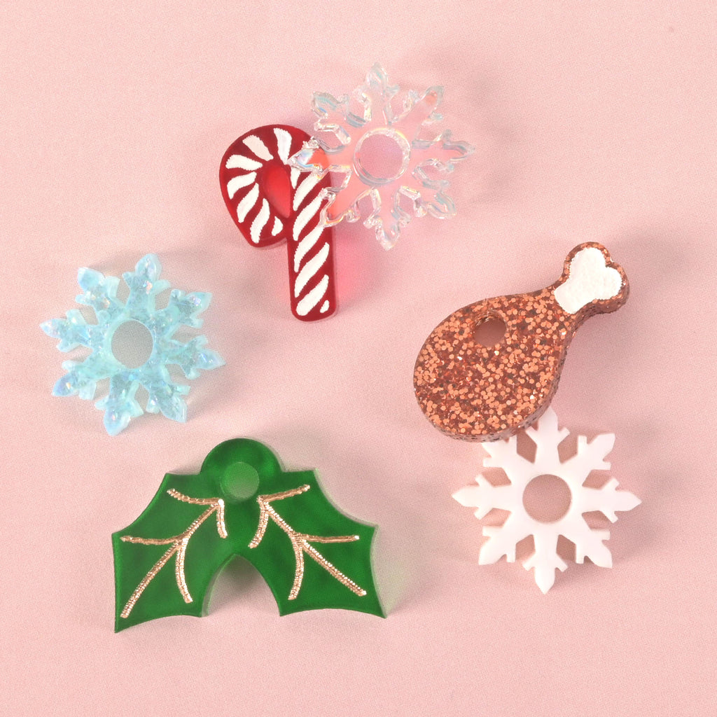 A collection of assorted holiday charms and pins on a solid pink background. The items include a red and white candy cane, a glittery copper drumstick, a clear iridescent snowflake, a blue iridescent snowflake, a white snowflake, and a green holly leaf charm with gold details.