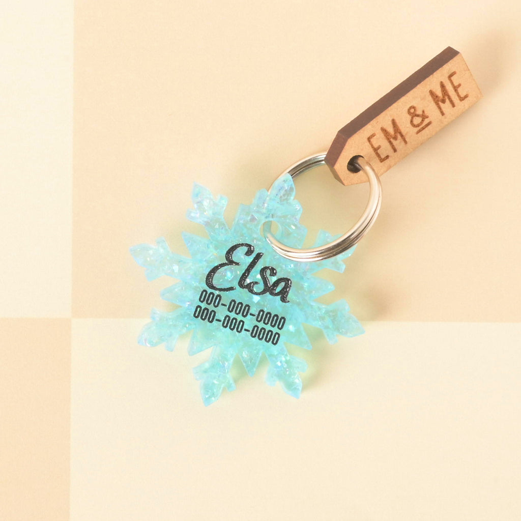 Blue glitter snowflake-shaped pet ID tag engraved with the name Elsa and phone numbers EM & ME.