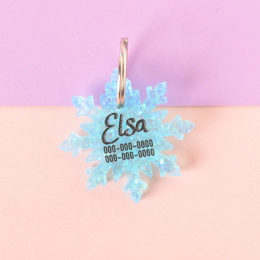 Blue glitter snowflake-shaped pet ID tag engraved with the name Elsa and phone numbers, displayed on a pastel pink and purple background.