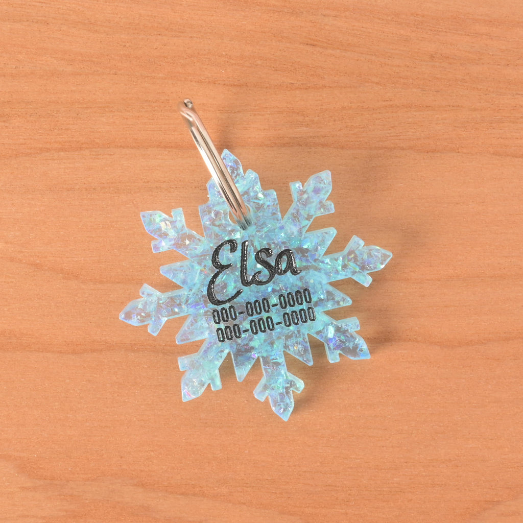 Close-up of a custom snowflake-shaped pet ID tag engraved with the name Elsa and a phone number, made from iridescent acrylic with a metal keyring.