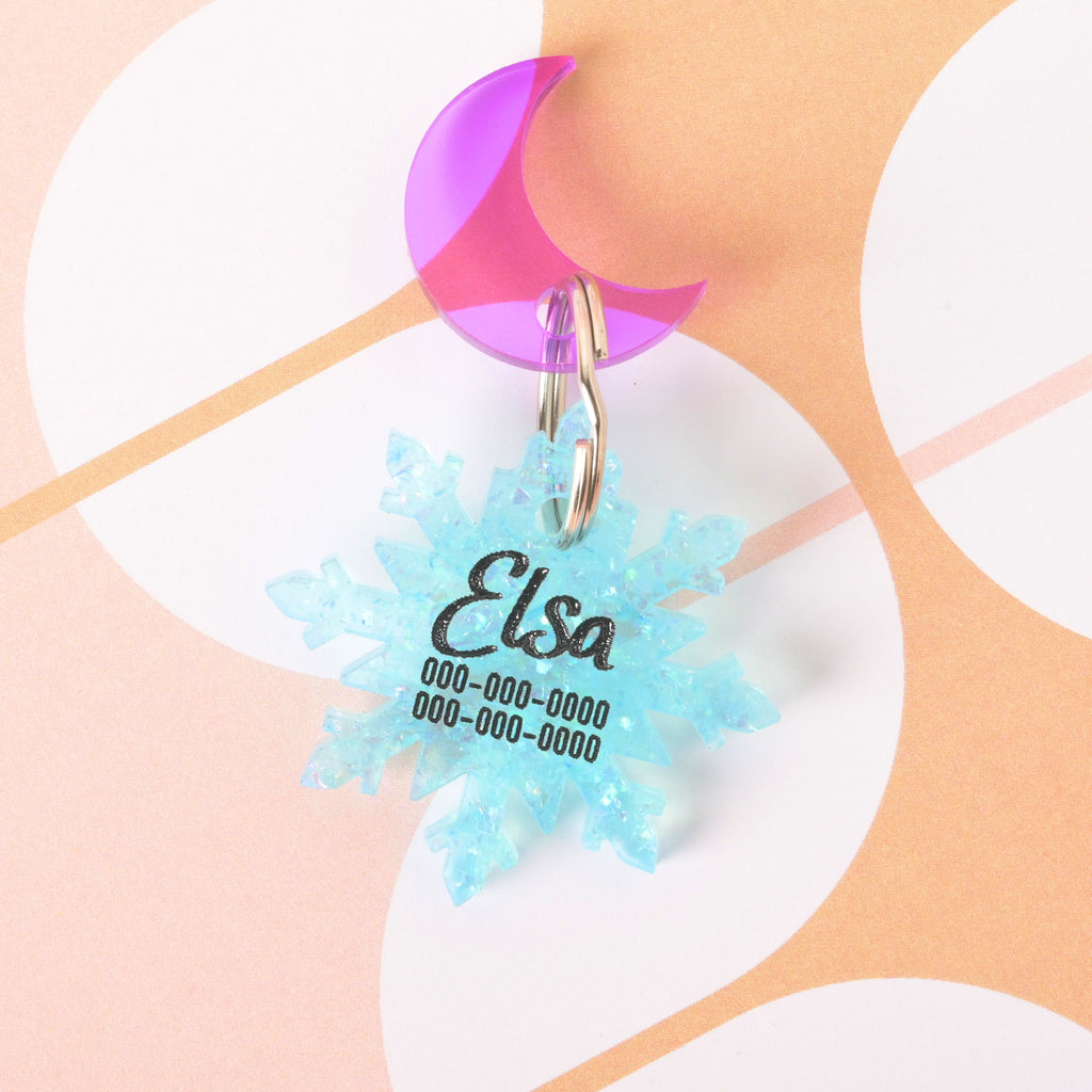 Blue glitter snowflake-shaped pet ID tag engraved with the name Elsa and phone number, paired with a purple acrylic crescent moon mini charm on a keyring, displayed on a patterned background.
