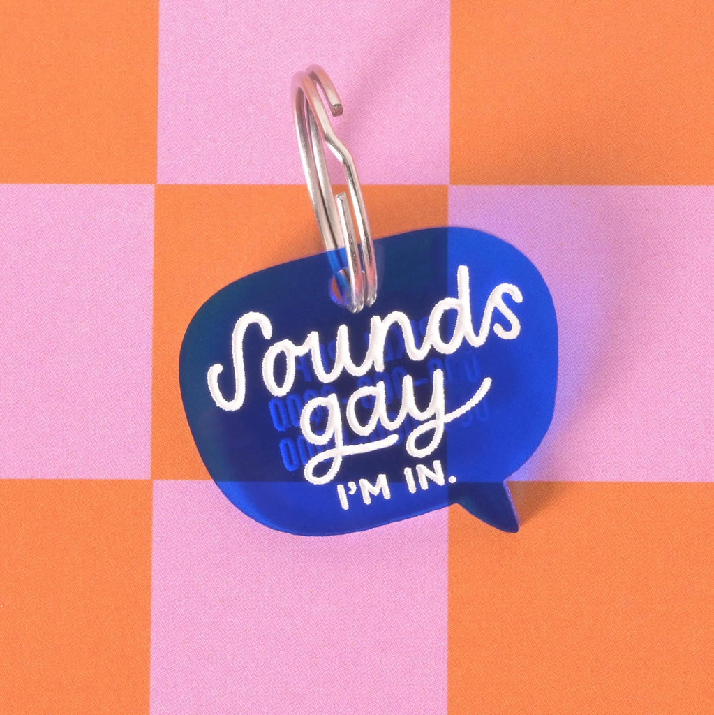 Close-up of a blue acrylic speech bubble dog tag with the phrase “Sounds gay, I’m in” in white text, displayed on a pink and orange checkerboard background.