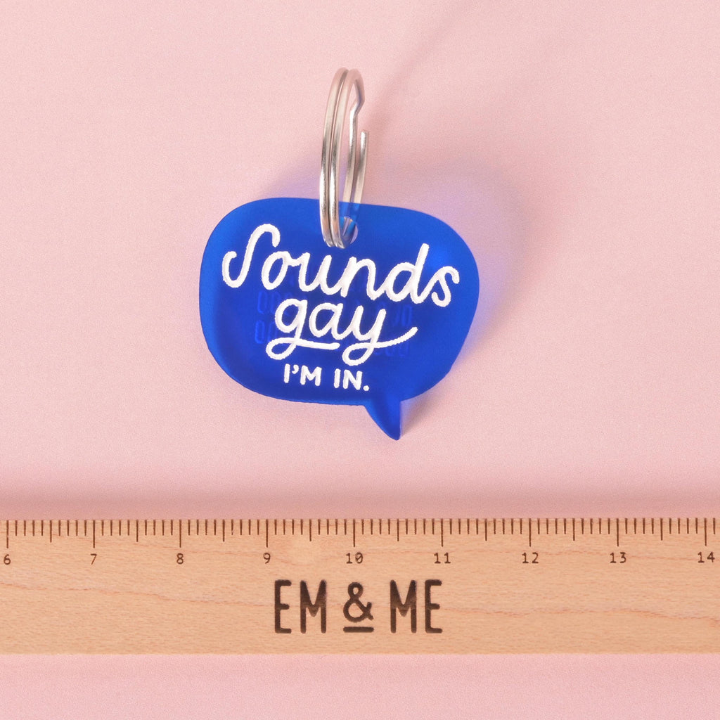 Blue acrylic speech bubble dog tag with the phrase “Sounds gay, I’m in” in white lettering, shown against a pink background with a wooden ruler for size reference.