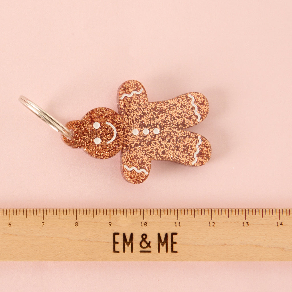 Close-up of a gingerbread man-shaped acrylic pet ID tag, featuring a brown glitter finish and white frosting details. The tag is displayed horizontally above a wooden ruler marked with the brand EM & ME, on a light pink background.