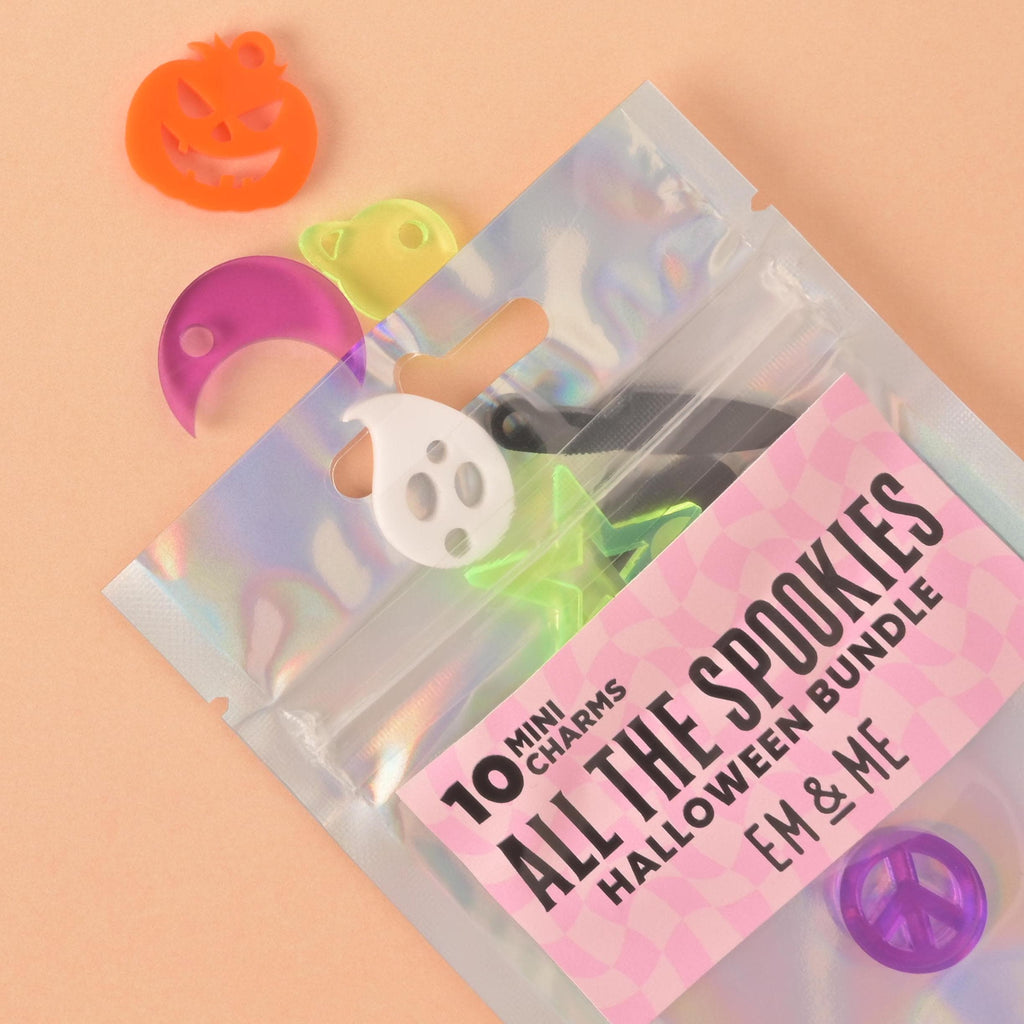 A clear, holographic bag filled with "spooky" themed charms. The bag is labeled "ALL THE SPOOKIES, HALLOWEEN BUNDLE" and "10 MINI CHARMS." Some charms are spilling out, including an orange jack-o'-lantern, a white ghost, a translucent green star, a purple crescent moon, a translucent yellow planet, and a purple peace sign. The background is a solid, light peach color.