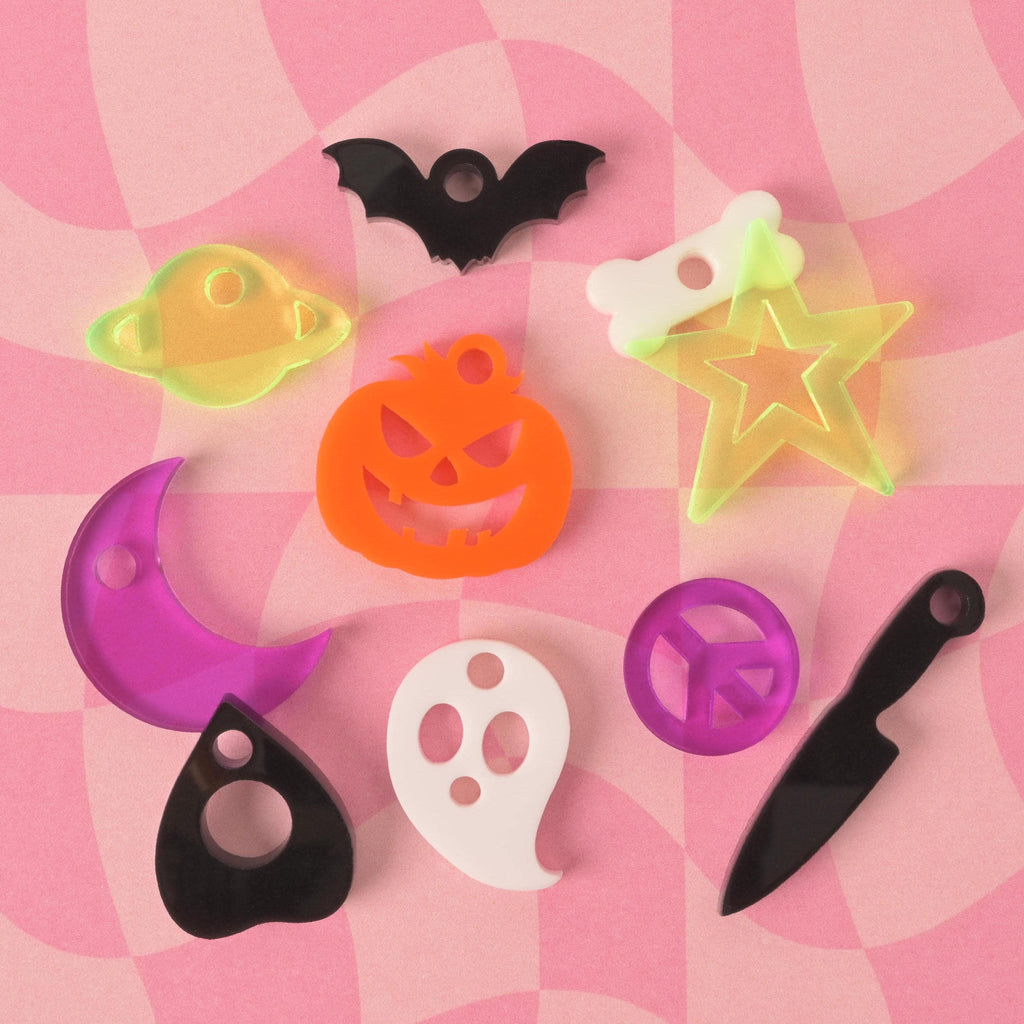 A collection of colorful, spooky-themed charms for pet tags, arranged on a pink and white checkered background. The charms include a black bat, a glow-in-the-dark star with a bone, a ghost, an orange jack-o'-lantern, a purple peace sign, a black knife, a purple crescent moon, a black planchette, and a translucent yellow-green planet with rings.