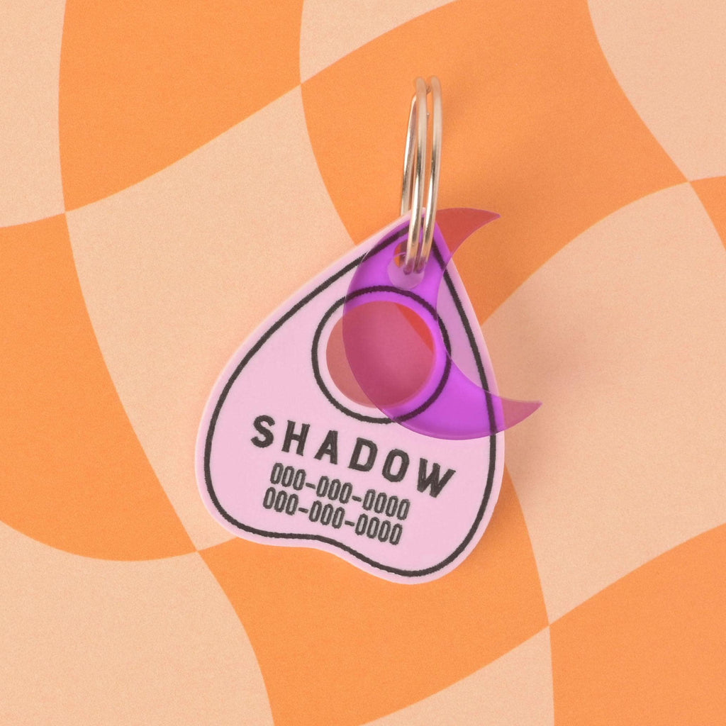 A personalized, pink, planchette-shaped pet ID tag with a black border. The tag is engraved with the name "SHADOW" and a phone number. A small, translucent purple moon charm is attached, partially overlapping the tag. Both are on a silver keyring and are shown on an orange and beige checkered background.