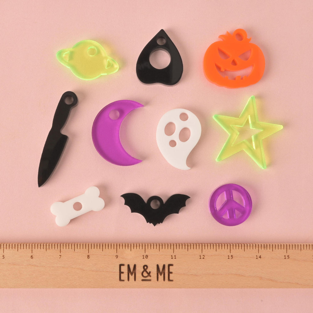 A flat lay of various spooky-themed pet charms on a pink background. The charms include a white bone, a black bat, a purple peace sign, a black knife, a purple crescent moon, a white ghost, a translucent green star, an orange jack-o'-lantern, a black planchette, and a translucent green planet. A wooden ruler with "EM & ME" is at the bottom of the photo.All The Spookies, Halloween Mini Bundle, Set of 10, Tiny Accessory for Pets, key chain, zipper pull or crafting, Add-on Charm