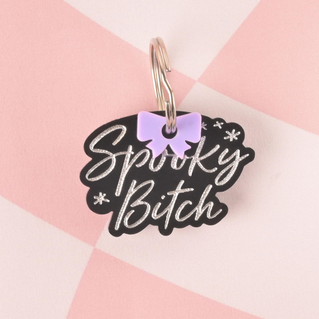 A black, personalized pet tag in a speech bubble shape that reads "Spooky Bitch" in a white script font. A small light purple bow charm is attached to the tag's ring. The item is displayed on a pink and white checkered background.