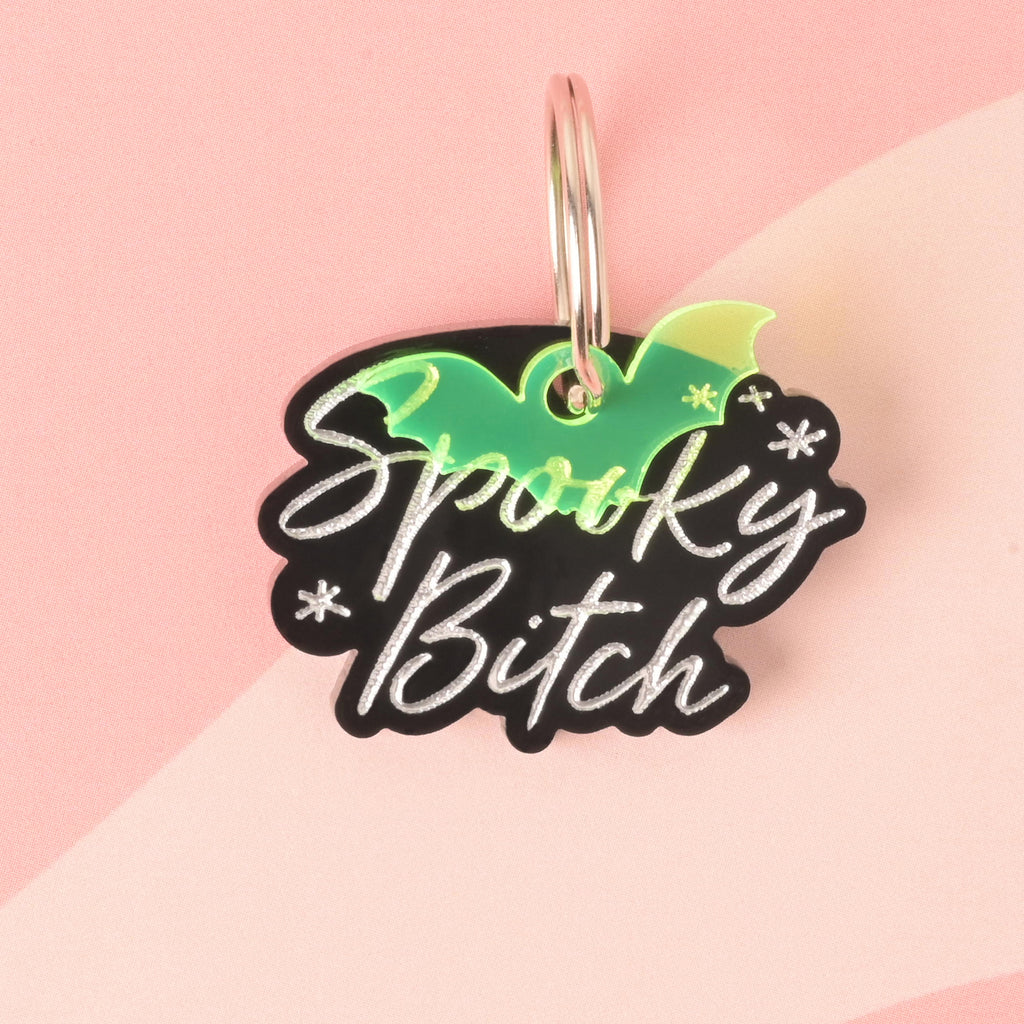 Black personalized pet ID tag that reads "spooky bitch" in silver and has a fluorescent green bat mini charm on solid pink background