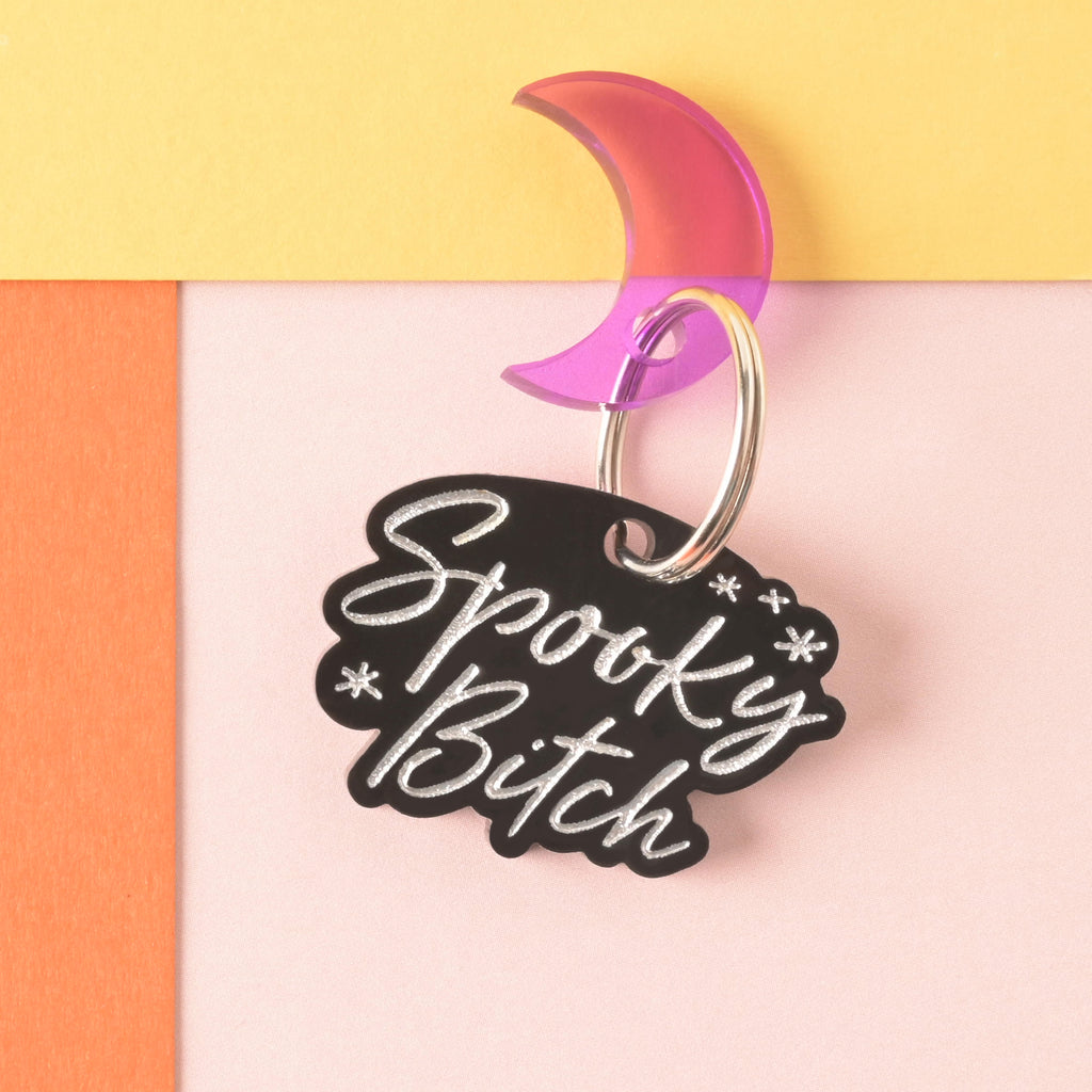 Black personalized pet tag that reads "spooky bitch" in silver script with a purple moon mini charm on color blocked background
