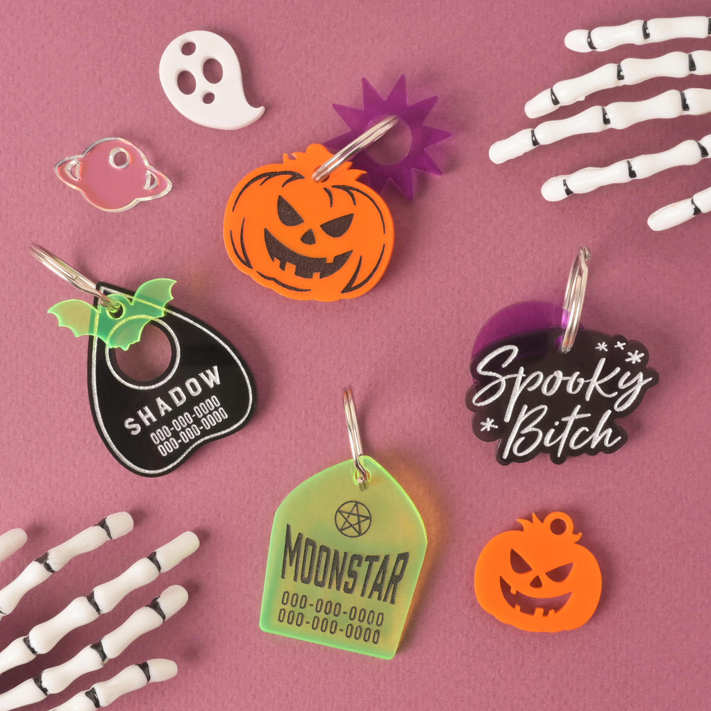 A flat lay photo of four custom pet ID tags and several small charms on a dark pink and purple background, with skeletal hands framing the edges. The tags include: a black planchette shape with green bat wings, engraved with "SHADOW"; an orange jack-o'-lantern shape with a purple starburst charm; a ghost-shaped tag that reads "Spooky Bitch"; and a green house or geometric shape engraved with "MOONSTAR" and a pentagram. A white ghost charm, a pink planet charm, and an orange jack-o'-lantern charm are also sc