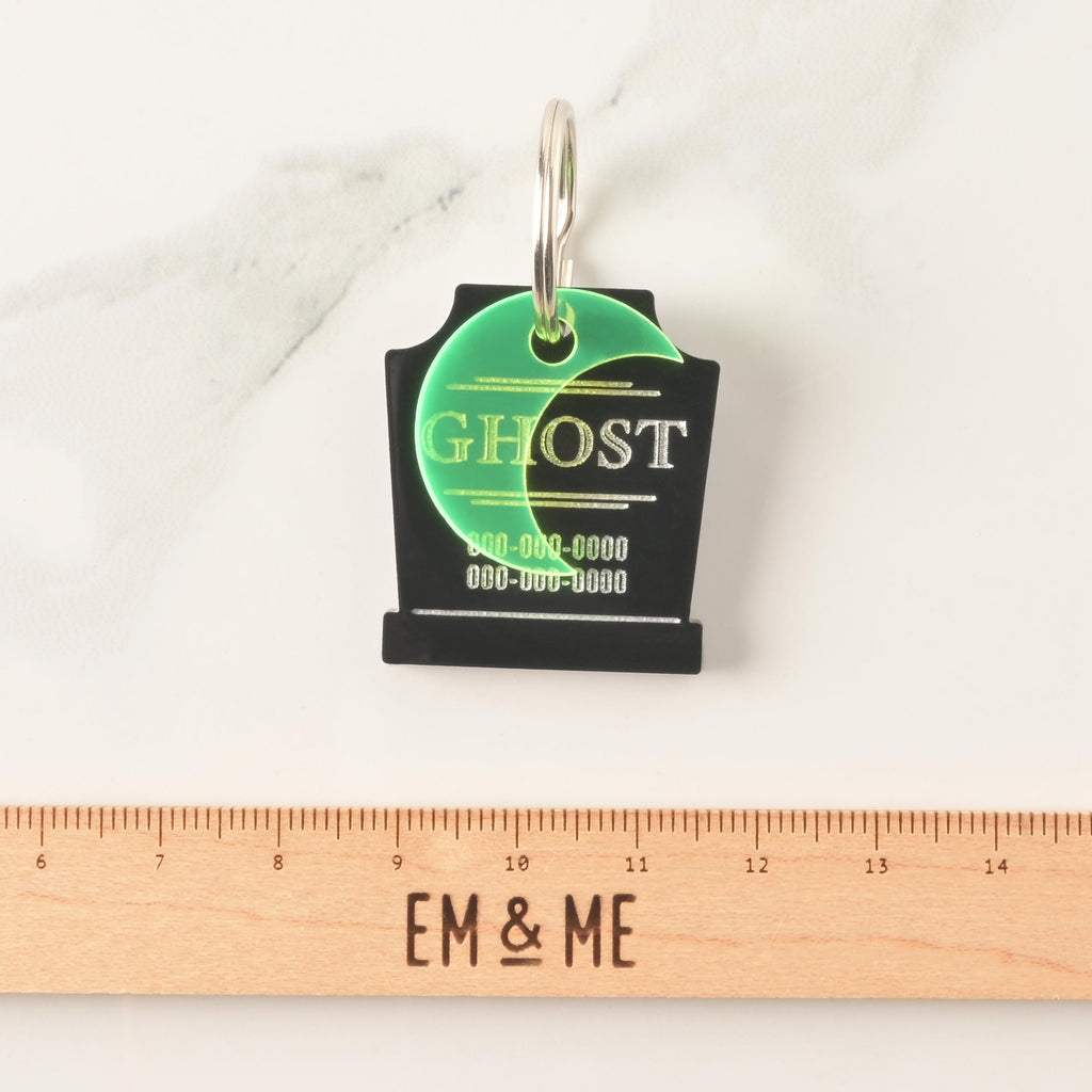 A close-up product photo of a black, tombstone-shaped pet ID tag. The tag is engraved with the word "GHOST" and two lines for phone numbers. A bright neon green crescent moon charm is attached to the top. A wooden ruler with the text "EM & ME" is positioned below the tag to show its size.