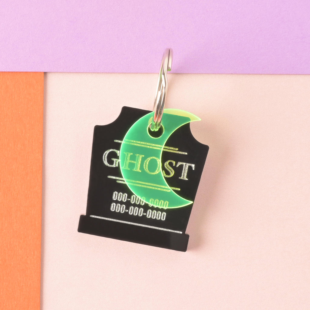 A close-up product photo of a black tombstone-shaped pet ID tag, engraved with the word "GHOST" and two lines for phone numbers. A bright neon green crescent moon charm is attached to the top of the tag, which is shown with a silver split key ring.