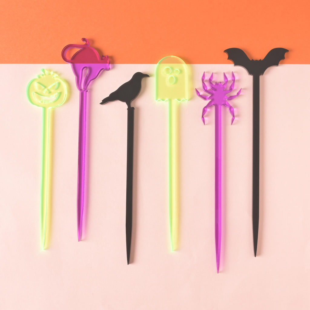 Set of 6 stir sticks, fluorescent green jack-o-lantern, purple cat, black crow, fluorescent green ghost, purple spider and black bat, on color blocked background
