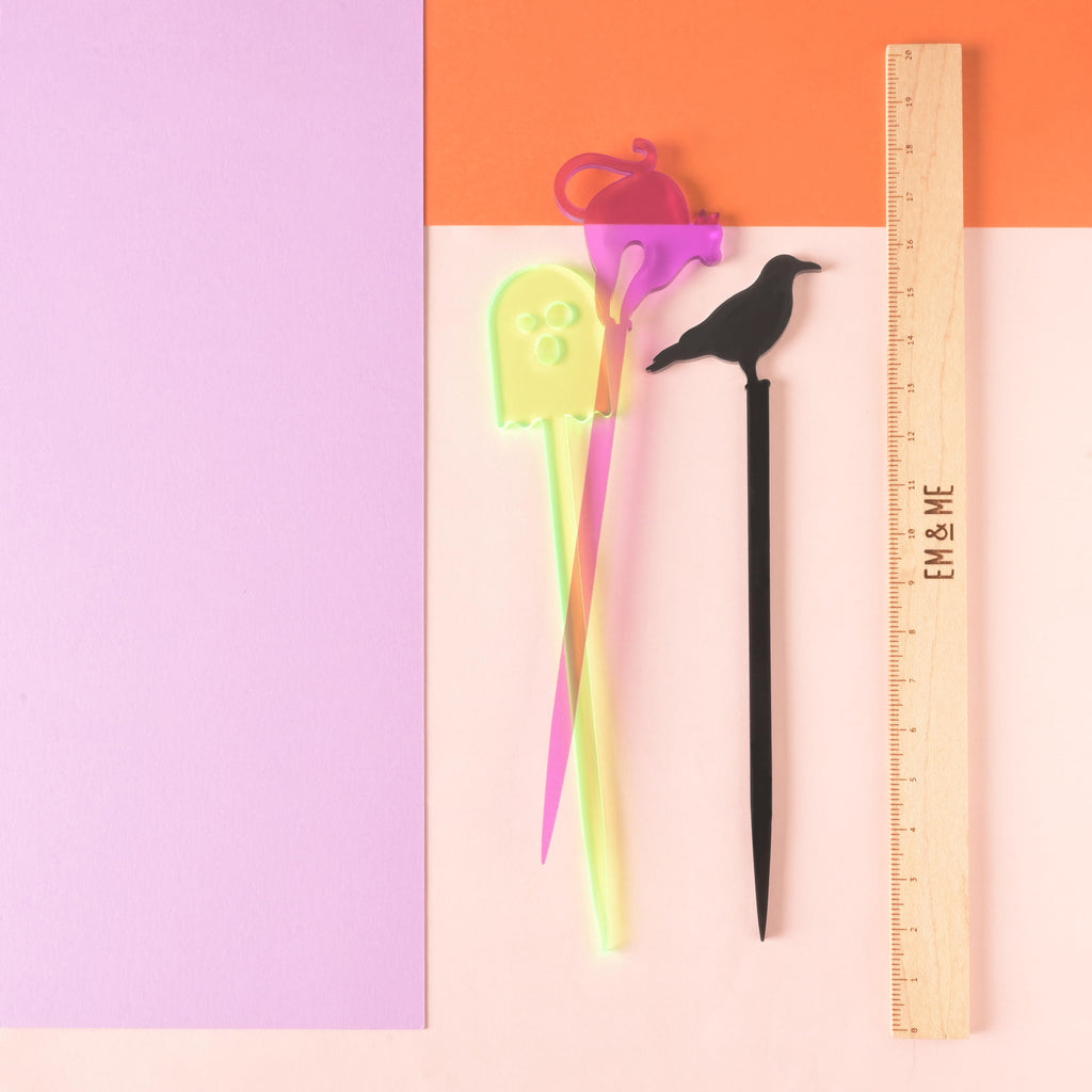 set of Three cocktail stir sticks, fluorescent green ghost, purple cat and black crow next to a wooden ruler that reads "EM & ME" on color blocked background