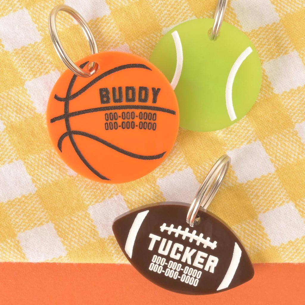 Set of three acrylic sports pet ID tags: an orange basketball engraved with Buddy, a green tennis ball, and a brown football engraved with Tucker, displayed on a yellow gingham background.