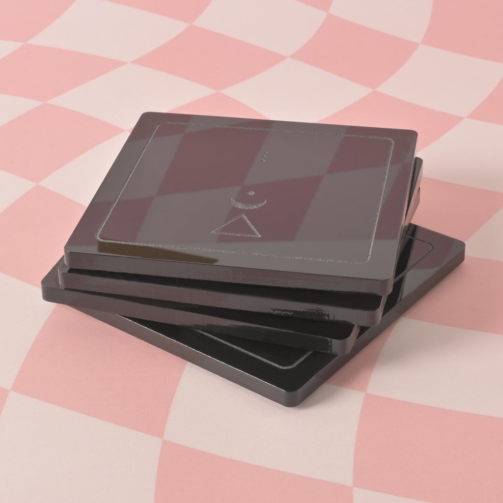 Alchemy Coasters, Set of 4, Minimal Astrology Symbols, engraved Coasters, A stack of black, square-shaped coasters is placed on a pink and white checkered background. A silver geometric design is visible on the top coaster.