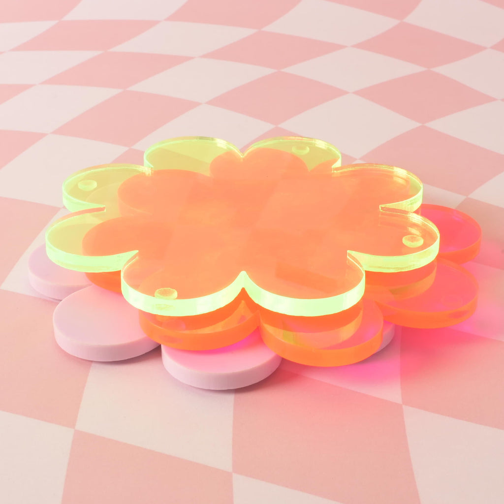 A stack of four flower-shaped coasters in different colors—fluorescent pink, fluorescent green, and lavender —is piled on a pink and white checkered background