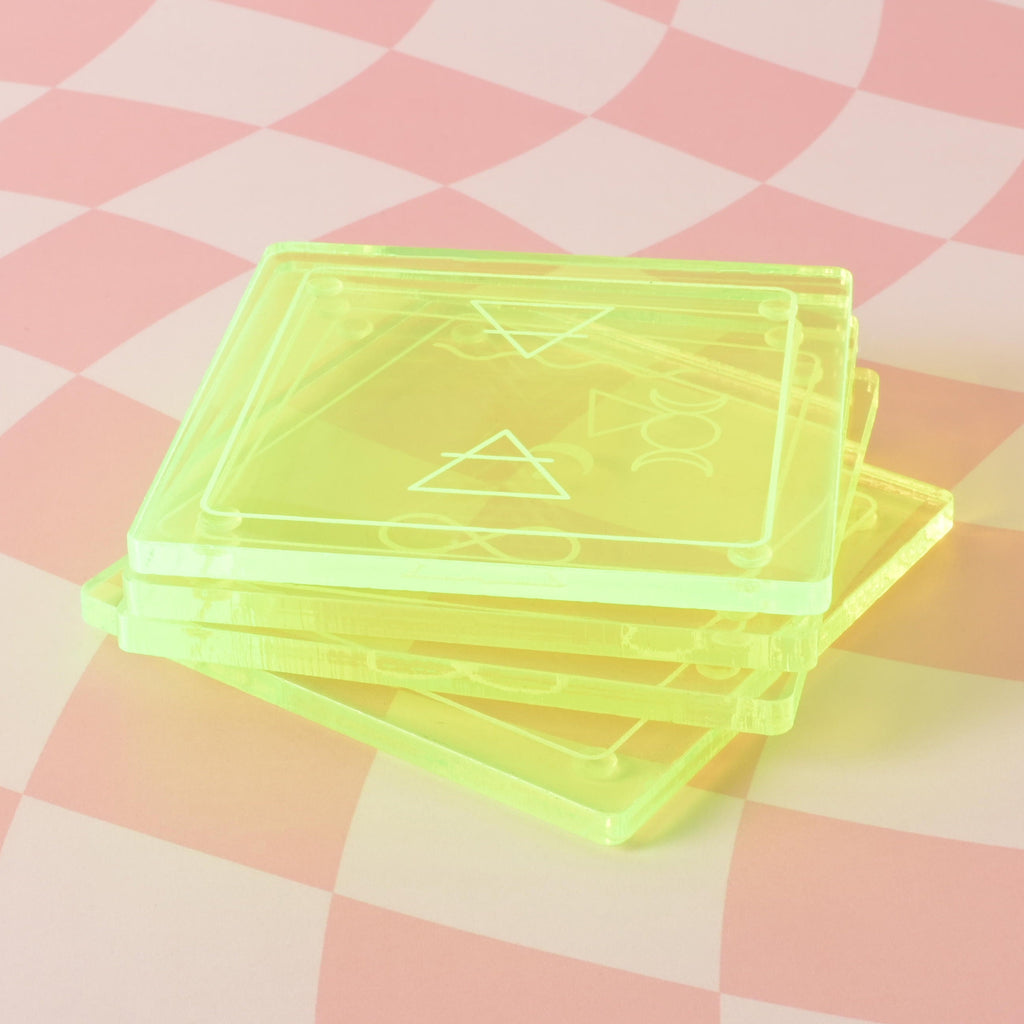 A stack of four square-shaped, translucent Fluorescent Green coasters is shown on a pink and white checkered background. Each coaster has a different etched design of a celestial or elemental symbol.