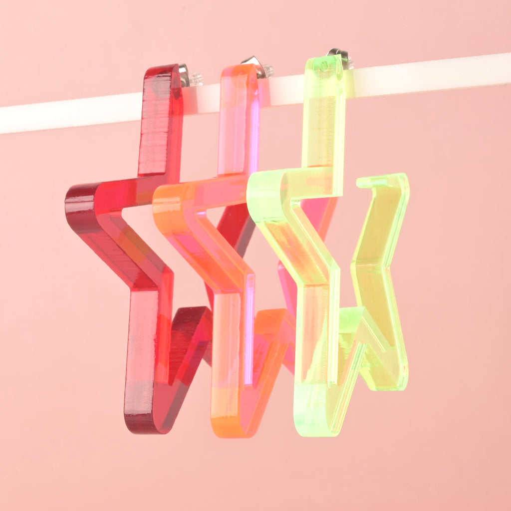 A set of three large, star-shaped hoop earrings in different colors hanging from a display bar. The colors include transparent red, fluorescent pink and red, and fluorescent green iridescent. The earrings are shown against a solid pink background.