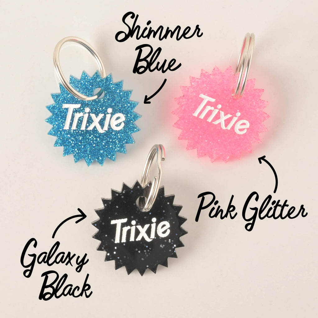 A product photo showing three starburst-shaped, glittery pet ID tags. The tag at the top left is blue glitter, labeled "Shimmer Blue." The tag on the top right is pink glitter. The tag at the bottom is black glitter, labeled "Galaxy Black." All three tags are engraved with the name "Trixie" and have a silver split key ring.