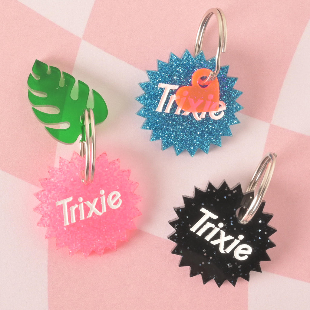 A product photo showing three starburst-shaped, glittery pet ID tags. The tag on the top right is blue glitter with a neon orange heart charm. The tag on the bottom left is pink glitter with a green monstera leaf charm. The tag on the bottom right is black glitter. All three tags are engraved with the name "Trixie" and have a silver split key ring.