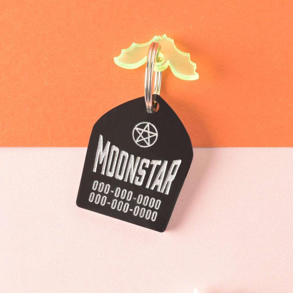 Opaque black tombstone-shaped personalized pet tag with fluorescent green bat mini charm. Tag has silver place holder text for pet name and phone numbers on color blocked background.
