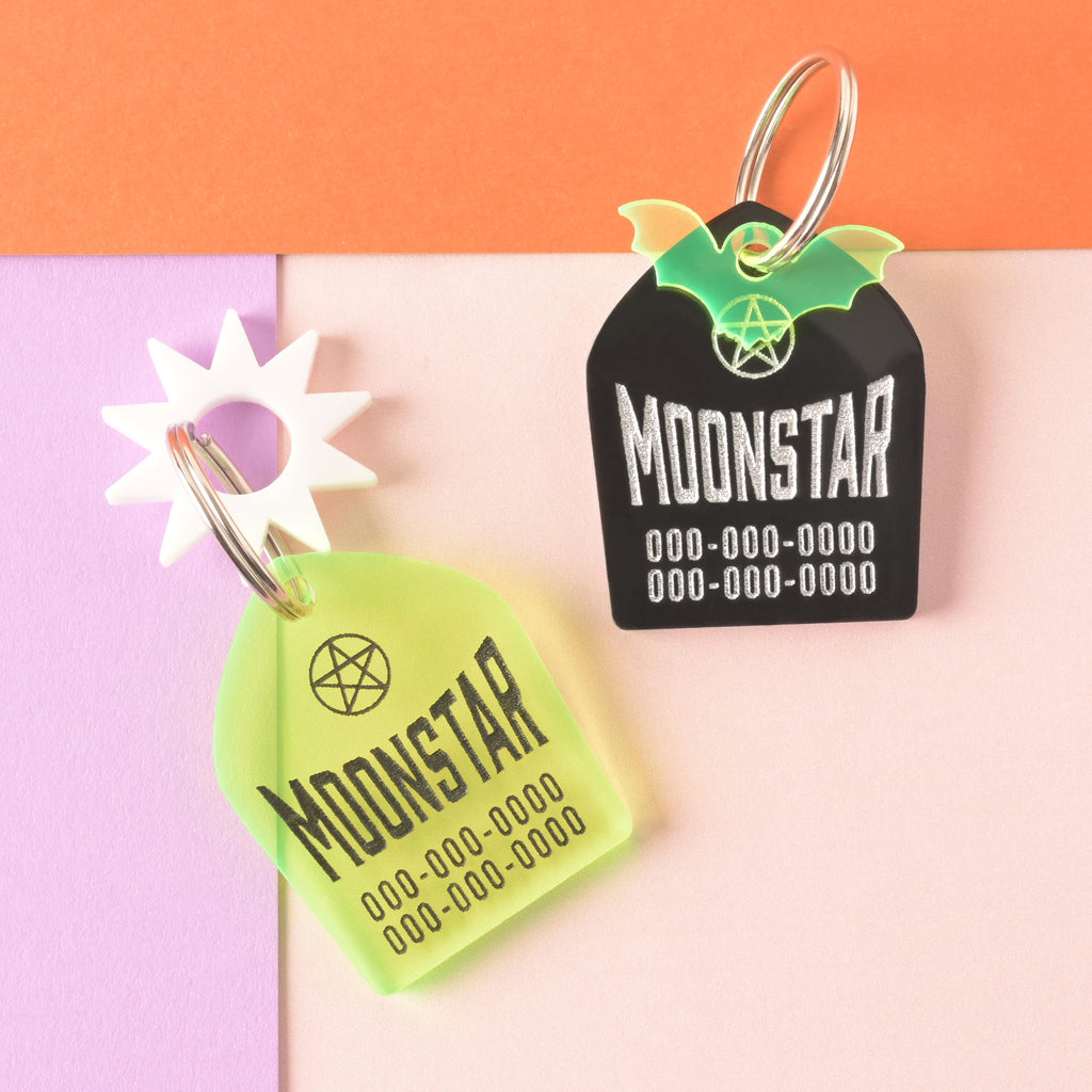 Set of two gravestone-shaped personalized pet tags. One fluorescent green with a white sun mini charm, the other opaque black with a fluorescent green bat mini.